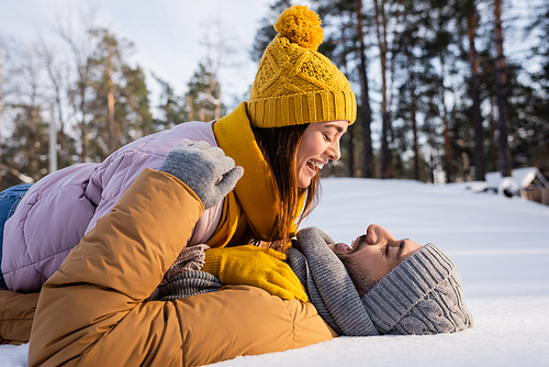 Side view of positive woman in knitted hat lying on boyfriend on snow in park