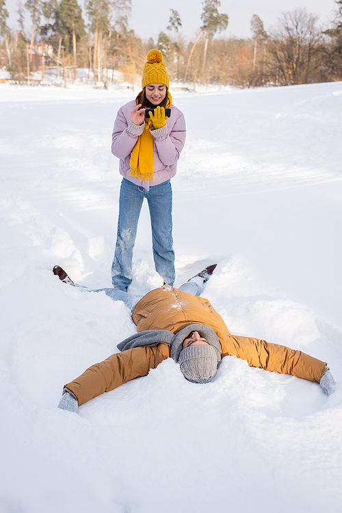 Smiling woman taking photo while boyfriend making snow angel in park