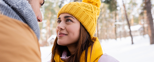 Young woman in knitted hat looking at boyfriend in winter park, banner