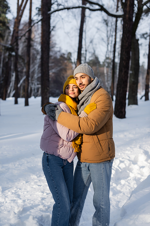Young couple in winter outfit embracing while standing in sunlight and snow