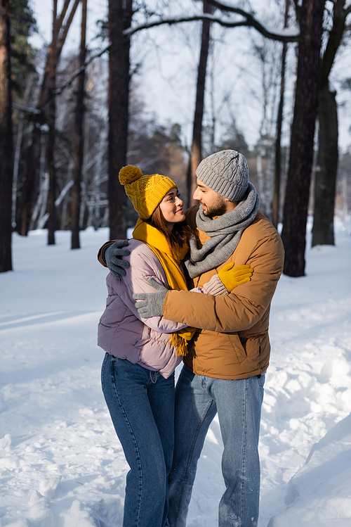 Smiling couple in warm clothes looking at each other while hugging in winter park