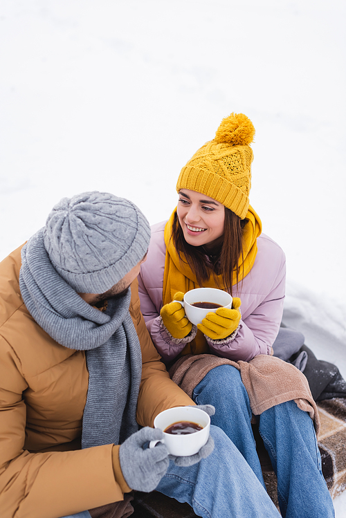 Smiling woman in knitted hat holding cup with coffee near boyfriend in winter park