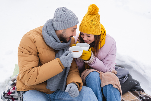 Positive couple holding cups of coffee sitting on blankets on snow