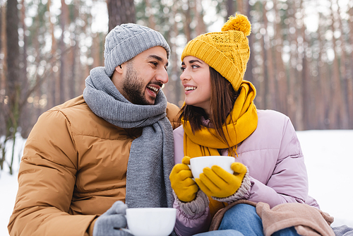Happy man in knitted hat holding cup near girlfriend in park during winter