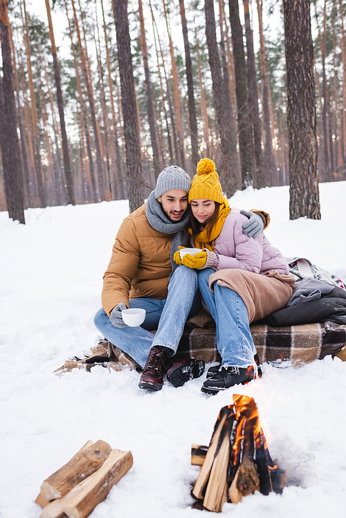 Man hugging girlfriend with cup near blurred bonfire on snow