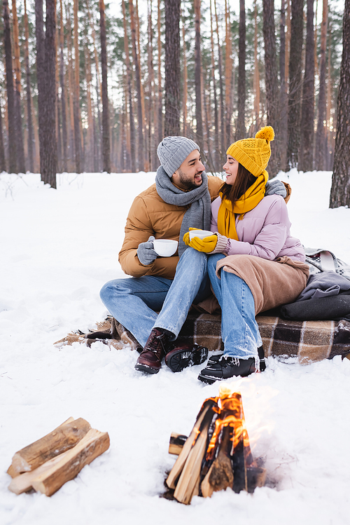 Happy young couple holding cups of coffee near bonfire and logs in winter park
