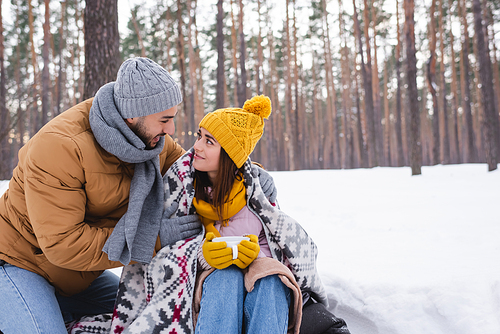 Happy man hugging pretty girlfriend in knitted hat and blanket holding cup in winter park