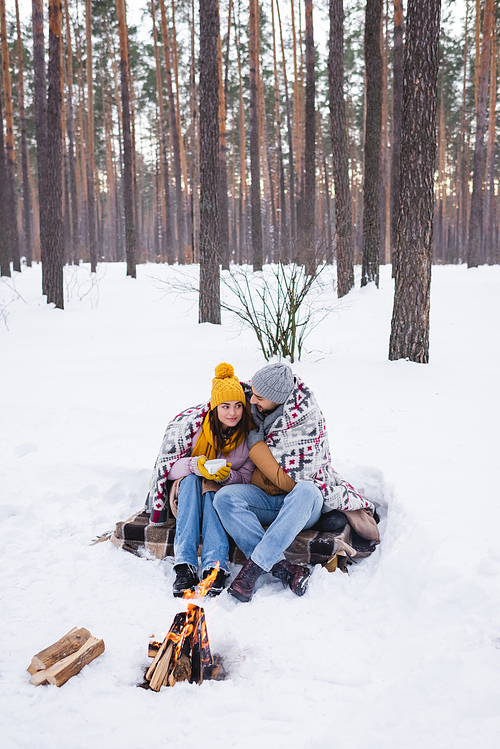 Young couple in blanket holding cup near bonfire in winter park