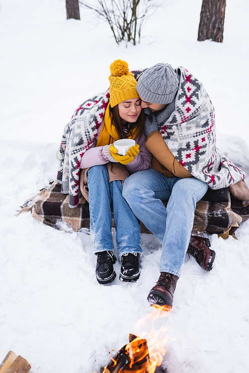 Young woman in winter outfit and blanket holding cup near boyfriend and bonfire on snow