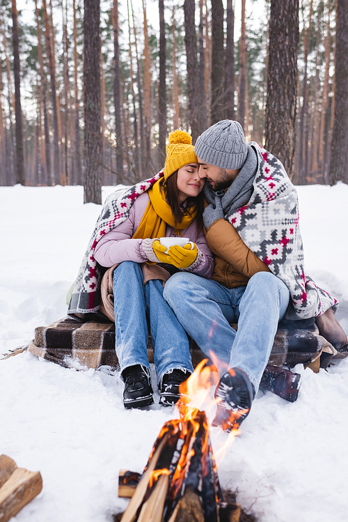 Young couple with blanket and cup warming near blurred bonfire in winter park