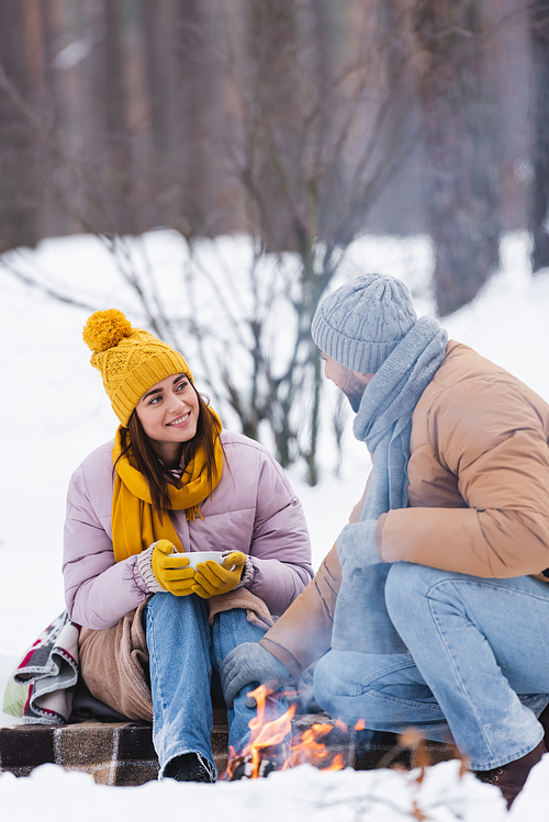 Smiling woman with cup looking at boyfriend near bonfire in winter park