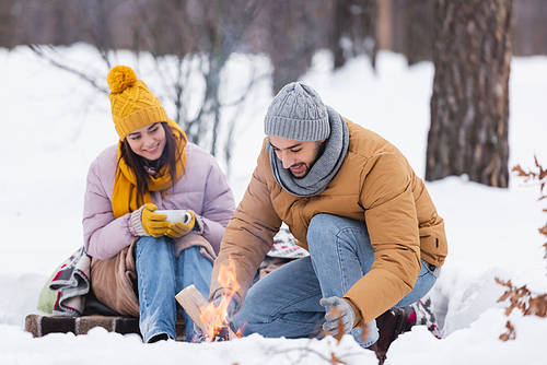 Cheerful man holding log near bonfire and girlfriend with cup in snowy park