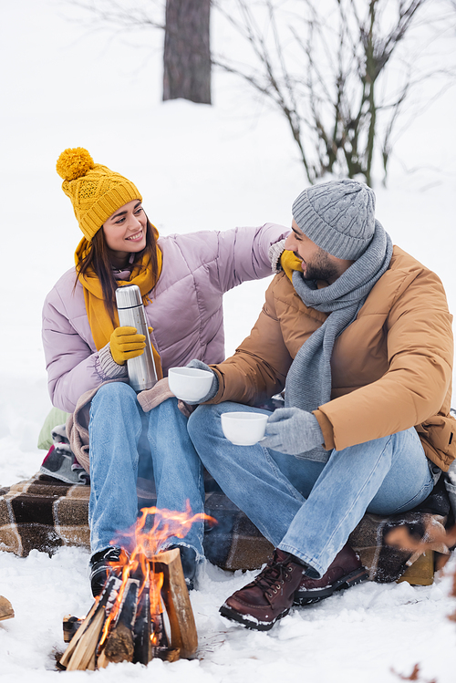 Cheerful woman holding thermos near boyfriend with cups and blurred bonfire in winter park