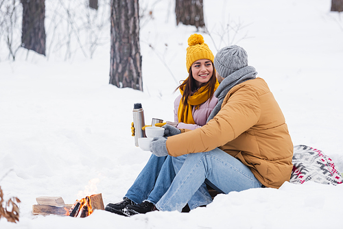 Man holding cups near smiling girlfriend with thermos and bonfire in snowy park
