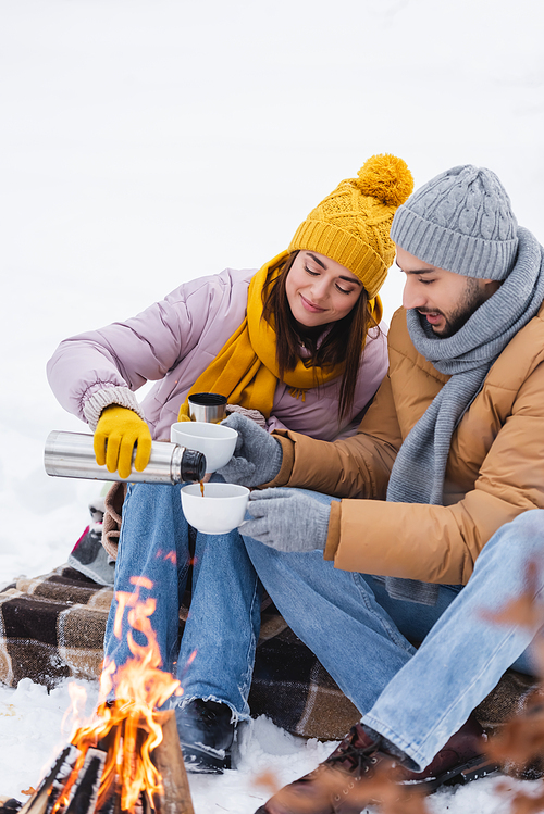 Man holding cups near girlfriend with thermos and blurred bonfire in winter park