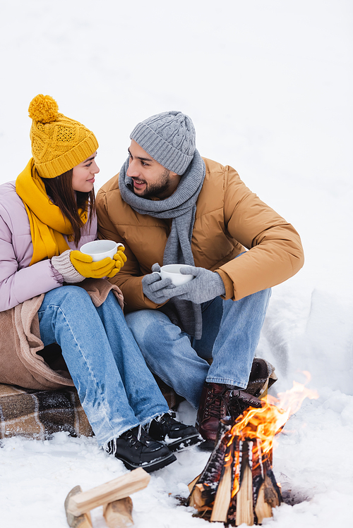Smiling man in gloves holding cup near girlfriend and bonfire on snow