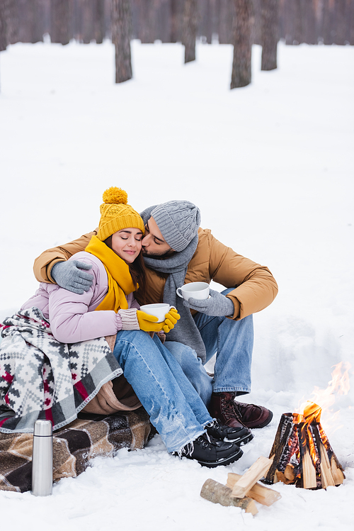 Man kissing girlfriend with cup and blanket near bonfire in winter park