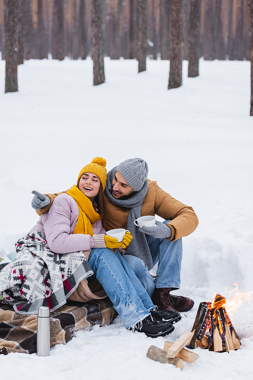 Smiling man pointing with finger near girlfriend with cup and bonfire in winter park