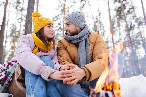 Young couple looking at each other while warming hands near blurred bonfire in winter park