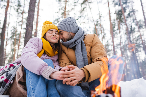Young couple sitting with closed eyes near blurred bonfire in winter park