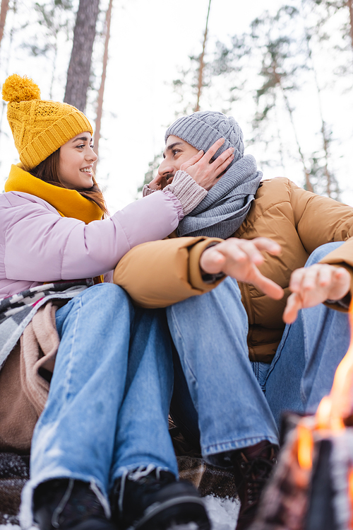 Low angle view of smiling woman adjusting knitted hat of boyfriend near blurred bonfire in winter park