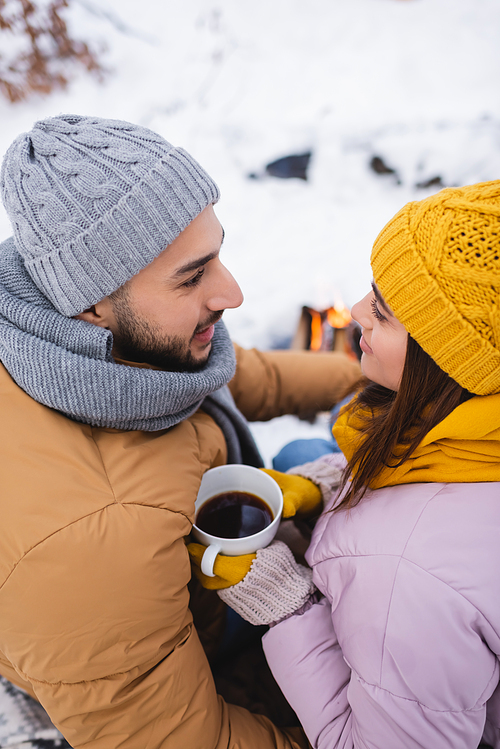 High angle view of woman in knitted hat holding cup of coffee near smiling boyfriend in winter park