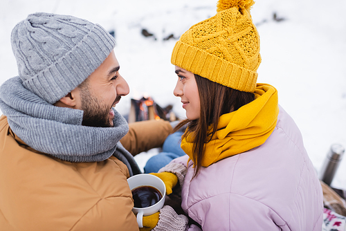 Side view of cheerful man looking at girlfriend with cup of coffee in winter park
