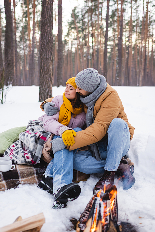 Man embracing girlfriend in blanket near cups and blurred bonfire