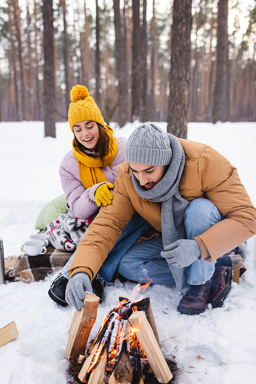 Smiling woman looking at boyfriend with wooden logs near bonfire in winter park