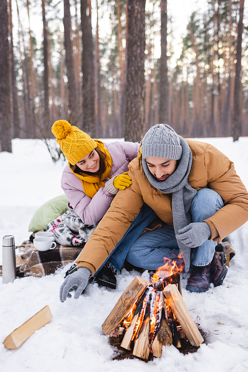 Smiling woman hugging boyfriend near cups, thermos and bonfire in winter park