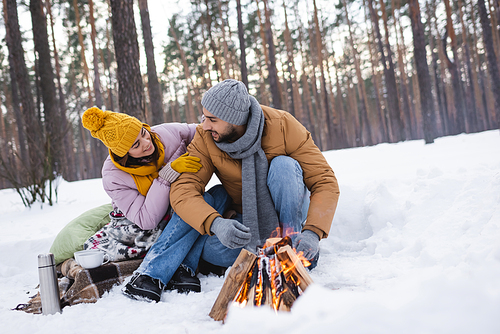Smiling woman looking at boyfriend in gloves near bonfire and cups in winter forest