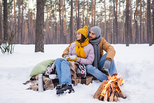 Young couple in winter outfit looking away near cups and bonfire in winter park
