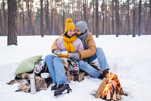 Man kissing girlfriend with cup near blankets and bonfire in winter park
