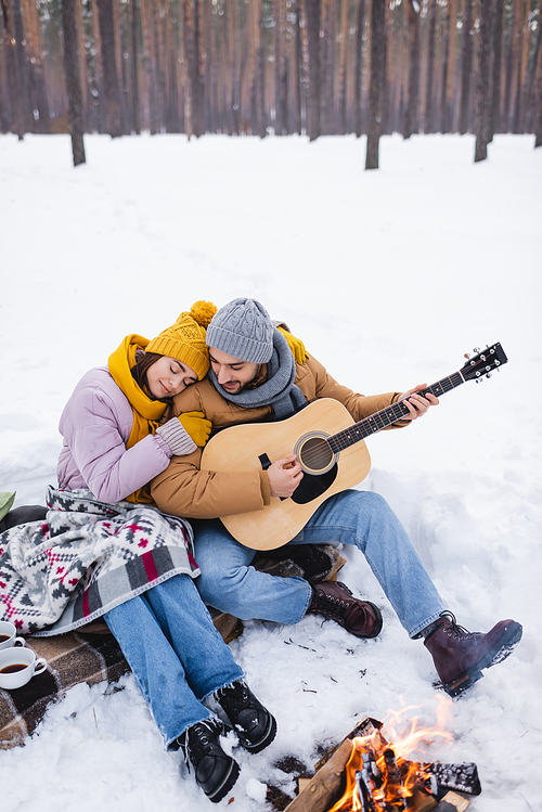 Smiling man playing acoustic guitar near girlfriend and bonfire in winter park