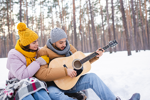 Smiling woman hugging boyfriend playing acoustic guitar in winter park