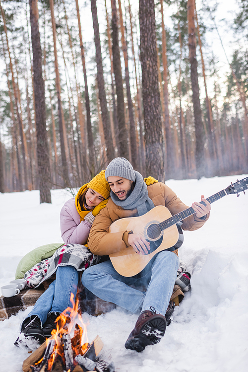 Man playing acoustic guitar near girlfriend and blurred bonfire in winter park