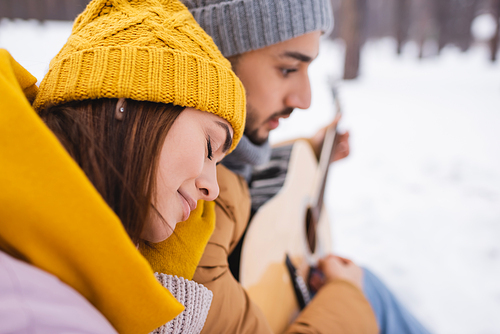 Side view of woman sitting near blurred boyfriend with acoustic guitar in winter park