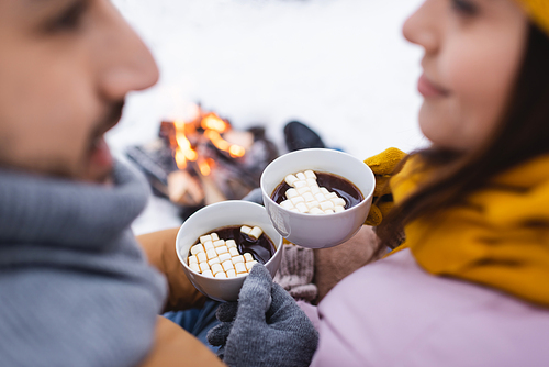 Cropped view of cups of cacao with marshmallows in hands of blurred couple in winter park