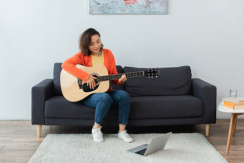 happy young woman learning how to play acoustic guitar near laptop on carpet