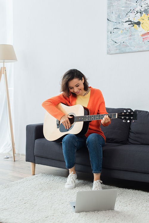 cheerful young woman learning how to play acoustic guitar near laptop on carpet