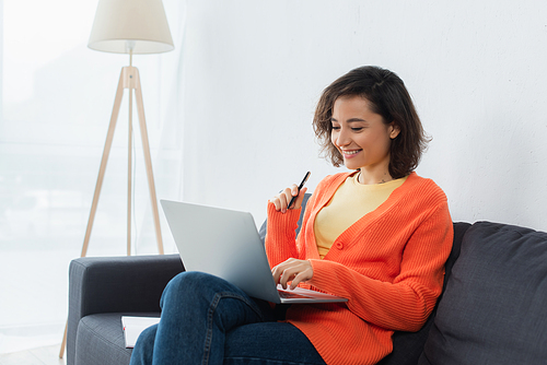 cheerful woman sitting on couch and holding pen while using laptop