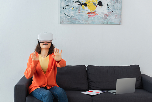 happy woman in vr headset gesturing while sitting near laptop on sofa