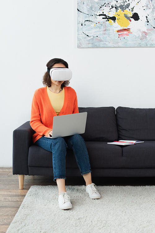 young brunette woman in vr headset using laptop while sitting on sofa