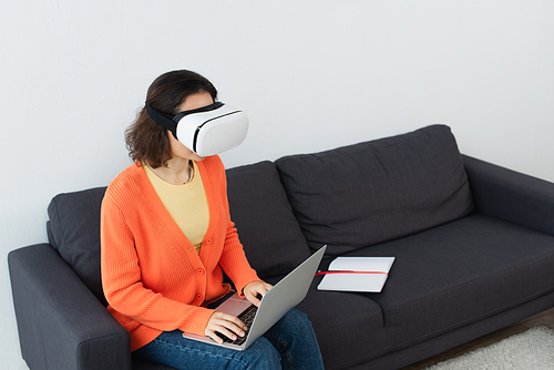 young brunette woman in vr headset using laptop while sitting on sofa