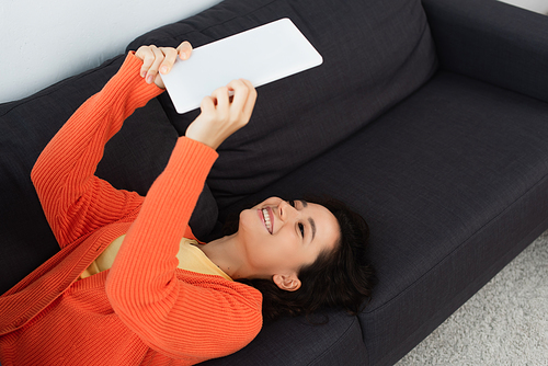 high angle view of happy young woman using digital tablet and lying on sofa