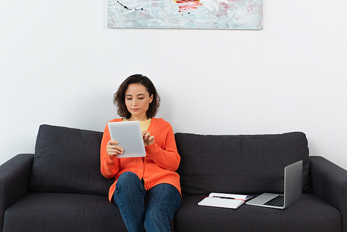 young woman using digital tablet and sitting near laptop and notebook on couch
