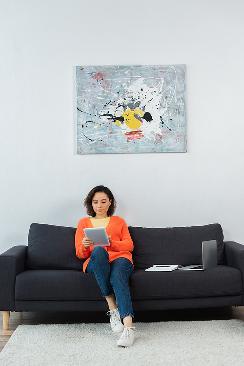young woman holding digital tablet and sitting near laptop and notebook on couch