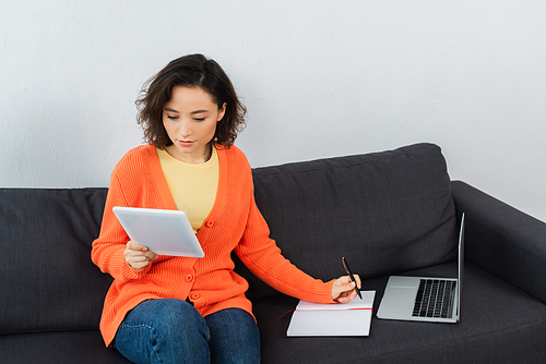 young woman looking at digital tablet and writing in notebook near laptop on couch