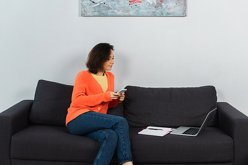 young freelancer holding smartphone and looking at laptop on couch