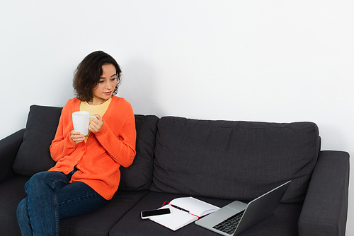 young freelancer holding cup of coffee near gadgets with blank screen on couch